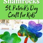 White and black text on green square on top half of image reads: Coffee Filter Shamrocks St. Patrick's Day Craft for Kids. Lower half of image shows one green and one rainbow tie dyed coffee filters cut into shamrock shapes in a bright wintery window.