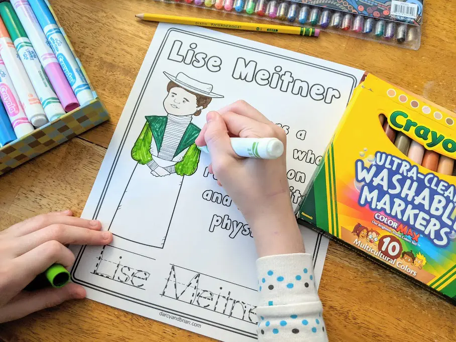 Overhead view of white girl's hand holding a marker and coloring the printable page for Lise Meitner. Markers and gel pens are on the table around the paper.