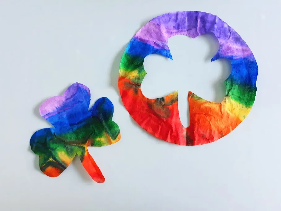 Rainbow colored coffee filter shamrock shape laying next to round filter with cut out in the middle.