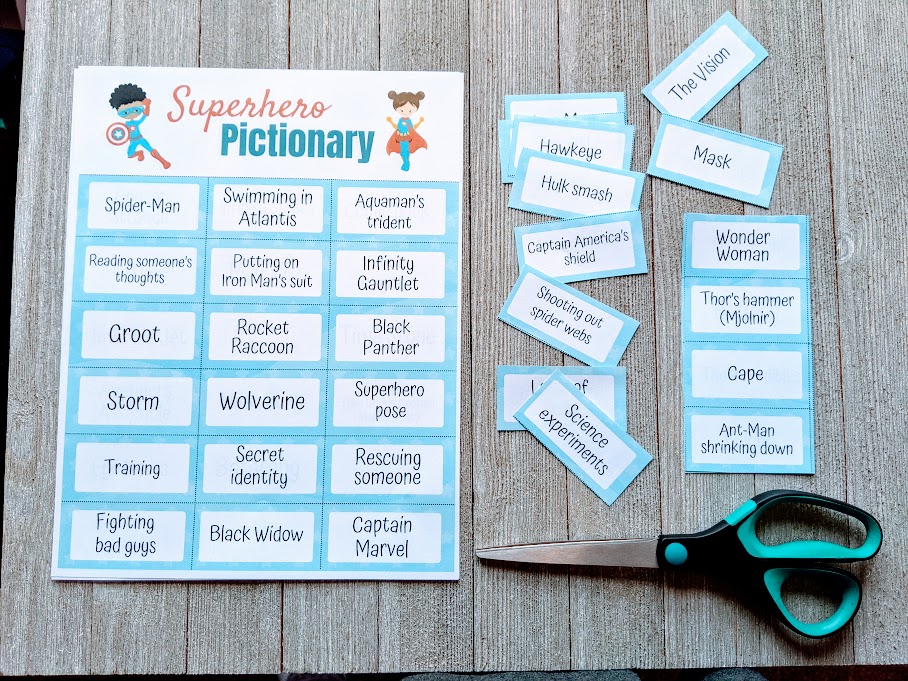 Overhead view of one full sheet of superhero pictionary prompts next to cut out clues and a pair of scissors on a dark gray wood background. 