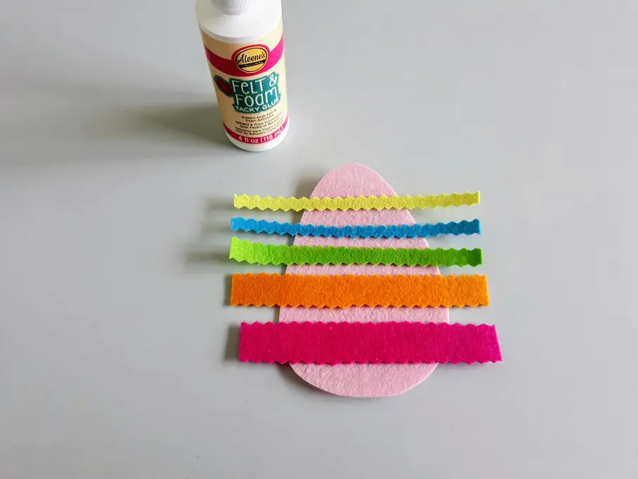 Bottle of felt glue next to pink felt egg shape with strips of felt glued across it.