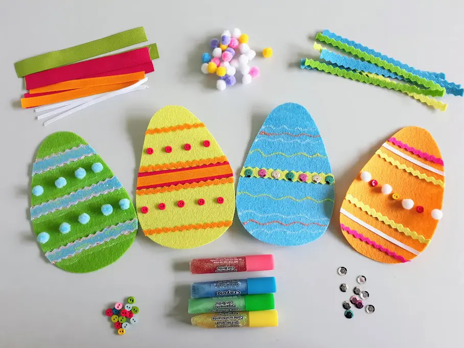 Overhead view of four completed felt Easter eggs with piles of different craft supplies around them such as felt, pom poms, sequins, and glitter glue.