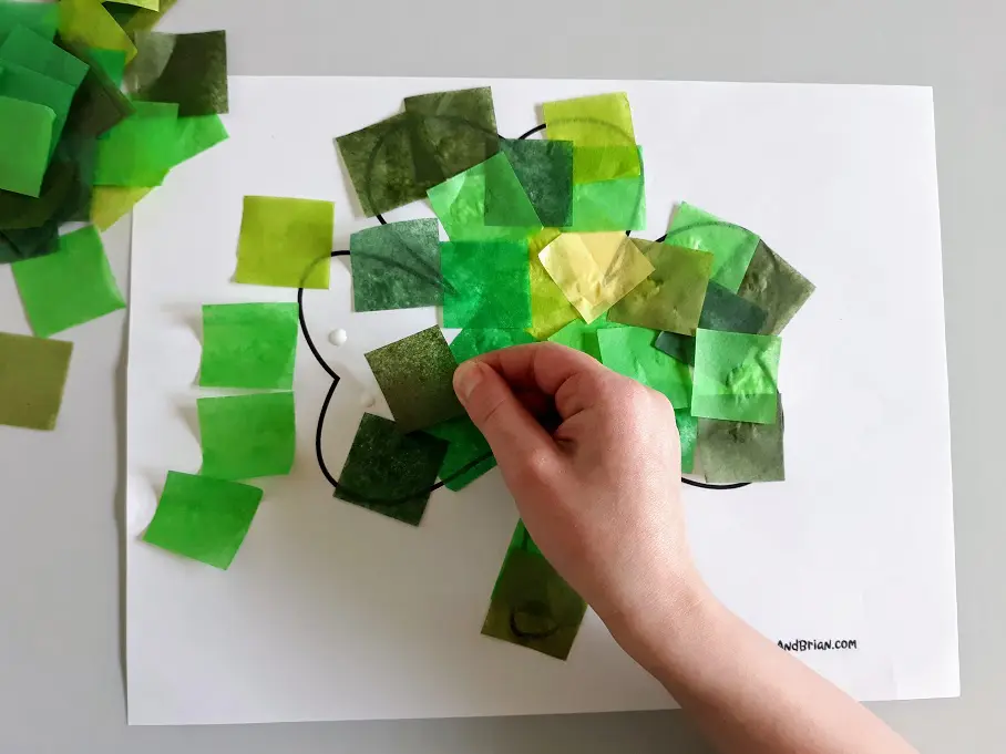 White child's hand gluing different shades of green tissue paper cut into squares onto printable shamrock template.
