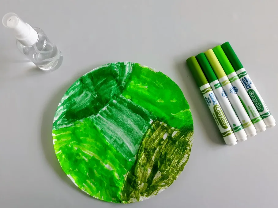 Round coffee filter colored with a variety of green markers. Markers and small spritz bottle set near filter paper.