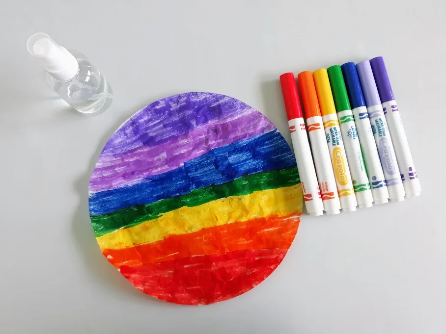 Overhead view of round coffee filter colored in a rainbow pattern. Markers and water spray bottle are next to the filter.