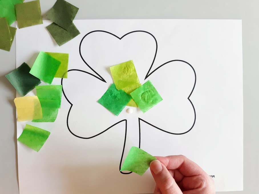 Pieces of cut up green tissue paper scattered on the left side of shamrock template. White child's hand gluing tissue paper to the shamrock.