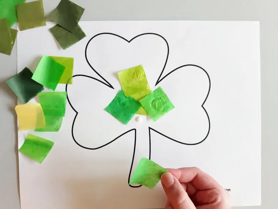 Pieces of cut up green tissue paper scattered on the left side of shamrock template. White child's hand gluing tissue paper to the shamrock.