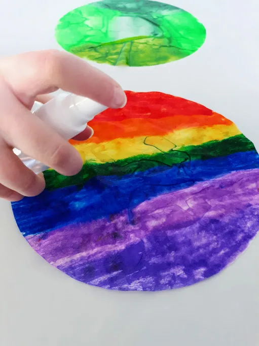 White girl's hand holding small spritz bottle and spraying rainbow colored coffee filter with water. A wet green colored coffee filter is farther in the back.