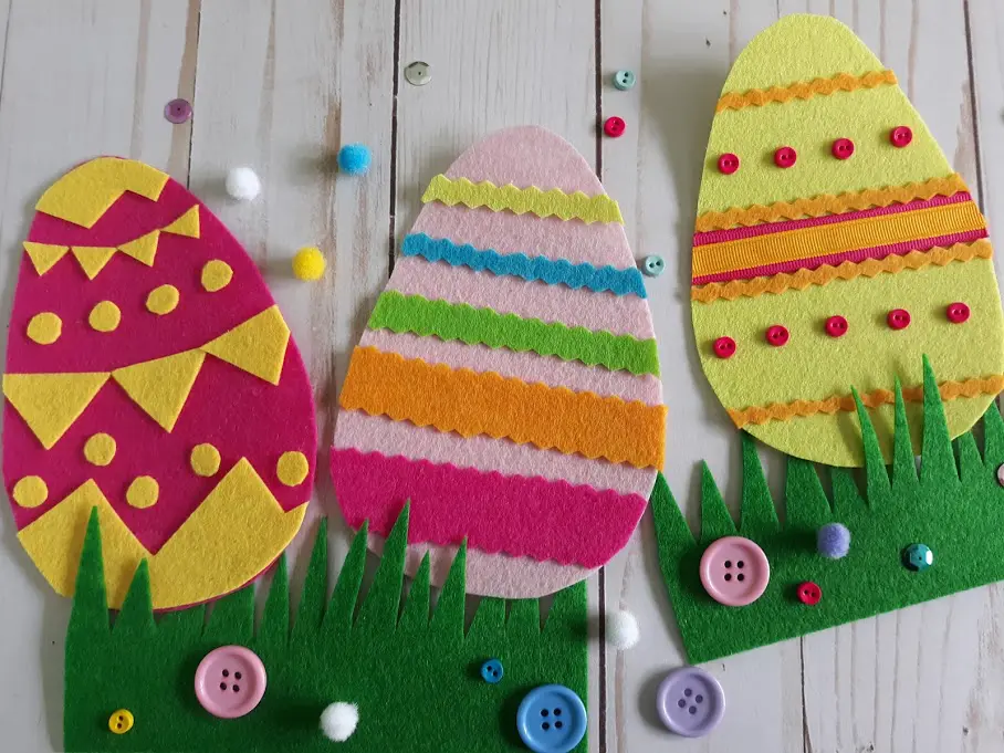 Overhead view of three completed felt Easter eggs decorated with felt and mini buttons and displayed in green felt cut to look like grass.