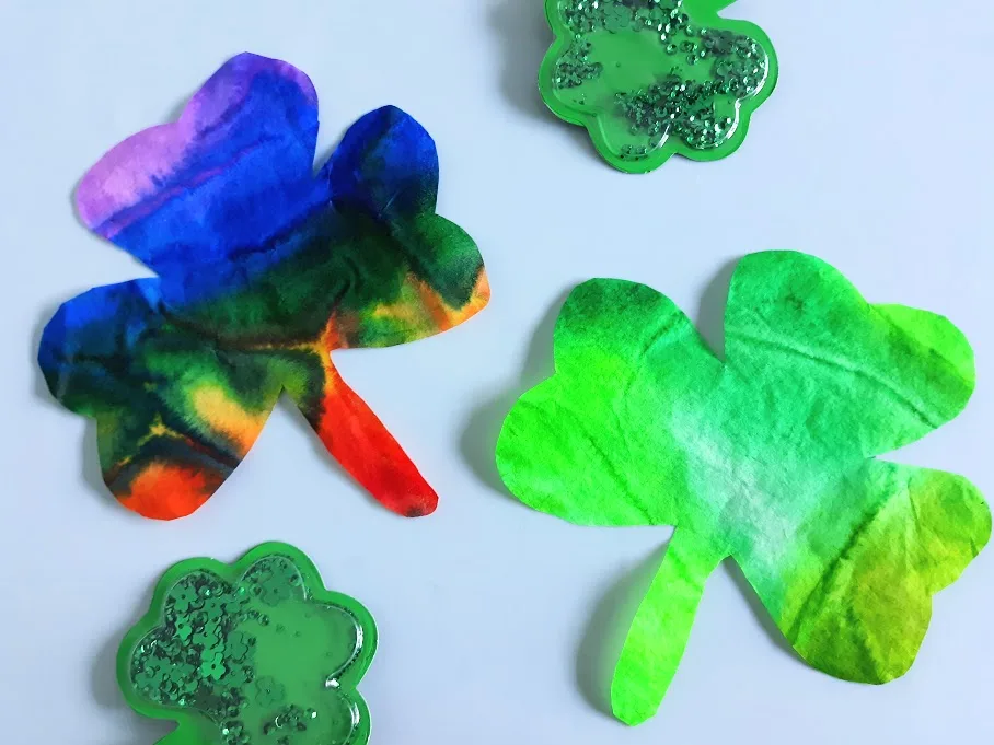 Finished rainbow and green coffee filter shamrocks laying on top of craft mat next to small plastic shamrocks.