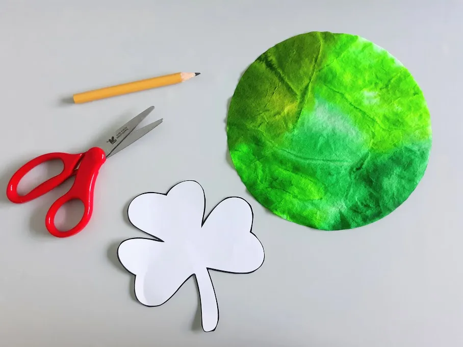 Overhead view of a pencil, red handle scissors, cut out shamrock template, and round green colored coffee filter.