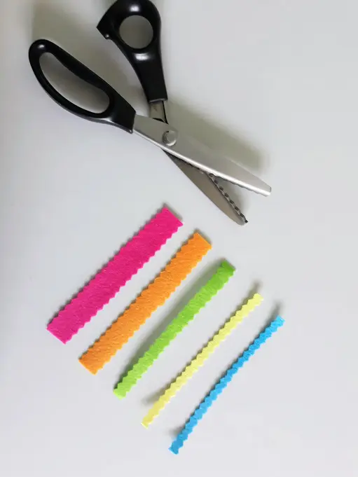 Overhead view of an open pair of serrated pinking shears and strips of felt cut with them to create zig zag edge.