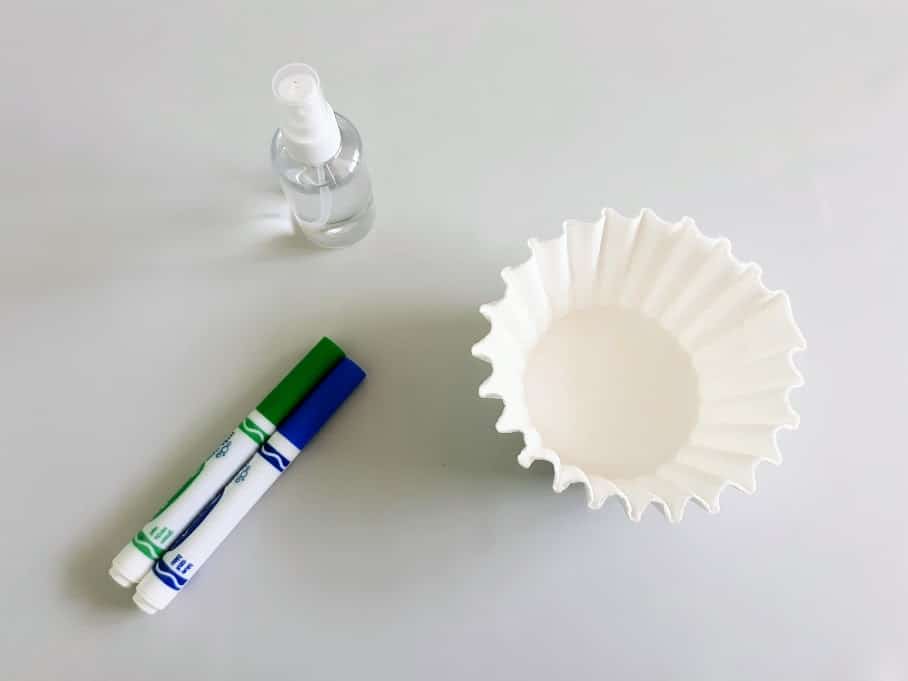 Overhead view of stack of white coffee filters, green and blue washable markers, and small spray bottle with water.