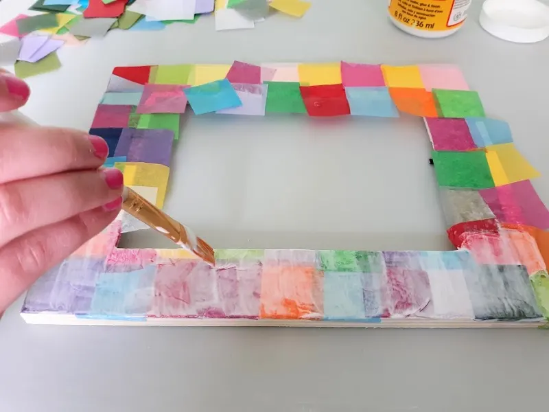 Side view of white girl's hand holding paint brush and applying mod podge coat over tissue paper applied to picture frame.