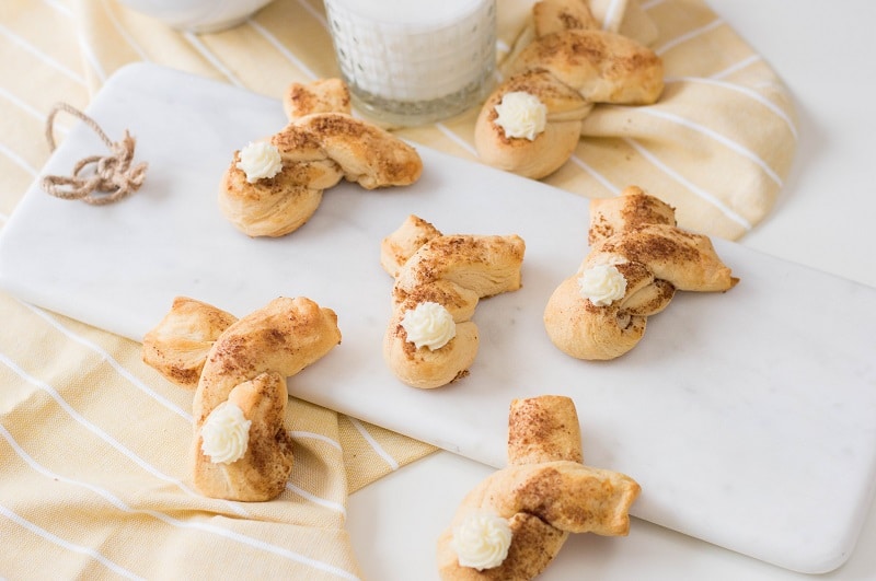 Six baked cinnamon twist bunnies with buttercream tails lay arranged over a small white cutting board and light yellow kitchen towels.