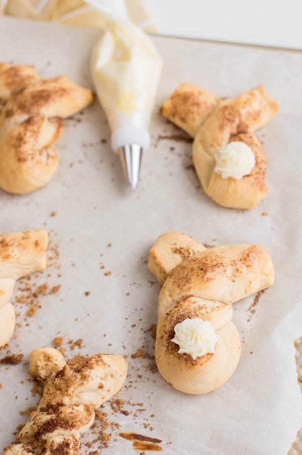 Overhead view of baked cinnamon twist bunnies on a baking sheet. Two have buttercream frosting tails. Piping bag full of frosting laying between decorated and undecorated bunnies.