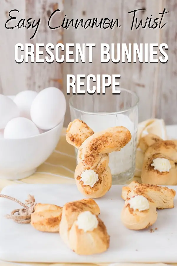 Three baked cinnamon twist bunnies laying across small white cutting board and table. One bunny shaped crescent roll propped up against glass of milk next to a small bowl of eggs. Top of picture has black and white text that reads: Easy Cinnamon Twist Crescent Bunnies Recipe.