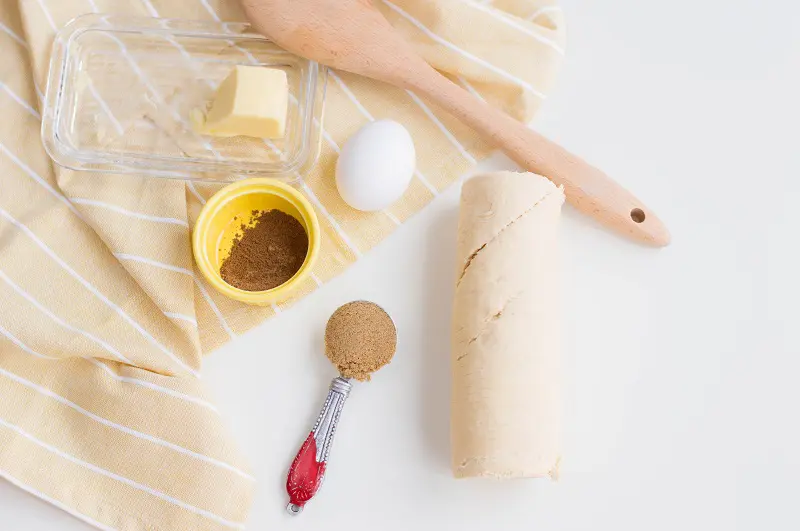 Unwrapped butter, one egg, small yellow bowl of cinnamon, measuring spoon full of brown sugar, roll of crescent dough removed from packaging, and wooden spoon arranged on yellow and white kitchen towel and white counter.