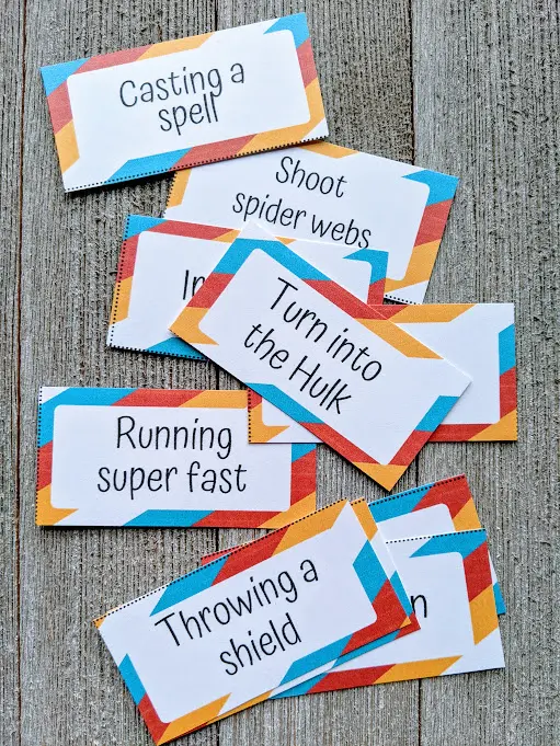 Close overhead view of about eight superhero charades clues cut out from the printable set. Each card has a red, orange, blue, and white border with the word prompt text in the middle. Visible clues include: casting a spell, turn into the Hulk, running super fast, and throwing a shield.