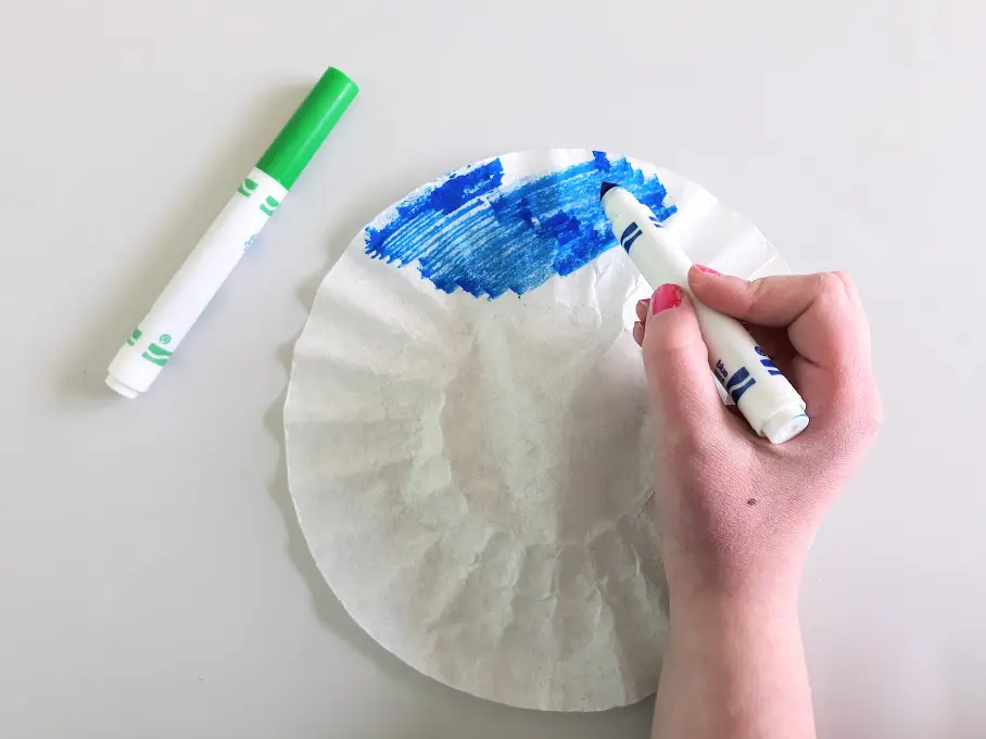 Overhead view of white girl's hand coloring coffee filter with blue washable marker. Green marker laying on the left side of the filter paper.