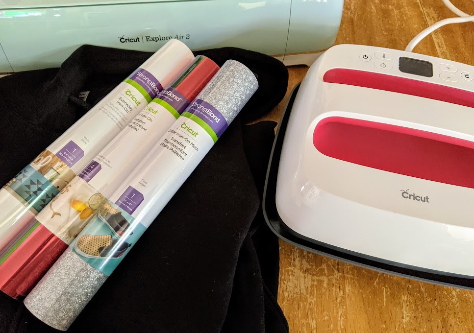 Supplies for project arranged on a table. Black hoodie folded up with three rolls of Cricut iron-on vinyls on top. Behind the hoodie is a Cricut Explore Air 2 machine and next to it is a Cricut EasyPress 2.