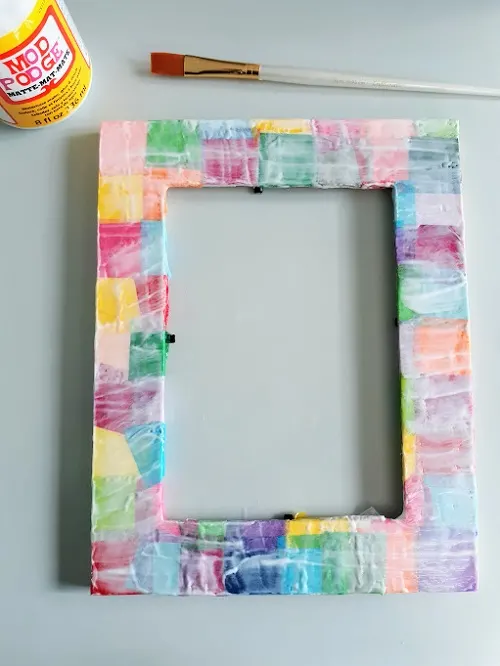 Overhead view of photo frame covered in tissue paper squares with edges fixed so none are hanging over, and covered in a coat of mod podge. A paint brush and bottle of mod podge are set down near top of frame.