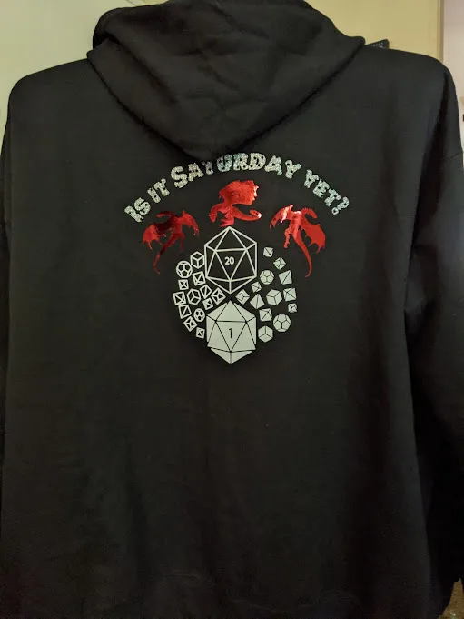 Back of black hoodie with Is it Saturday yet? in silver text above shiny red dragon silhouettes and black and white dice sets. Holding up finished custom hoodie project.