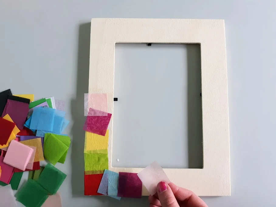 Overhead view of white girl's hand applying tissue paper square pieces to wood picture frame.