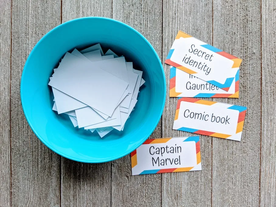 Overhead view of small round blue bowl with rectangle slips of paper inside. Next to the bowl are several pieces of paper with superhero theme words cut from printable charade word list.