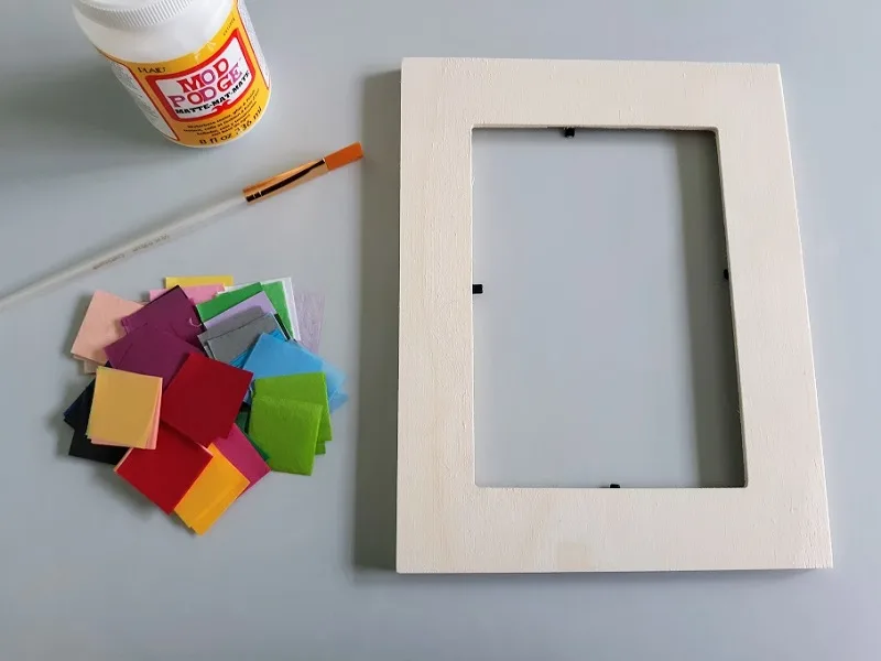 Overhead view of empty plain wooden picture frame laying on craft mat next to pile of pre-cut tissue paper squares in assorted colors, a paint brush, and a bottle of Mod Podge.