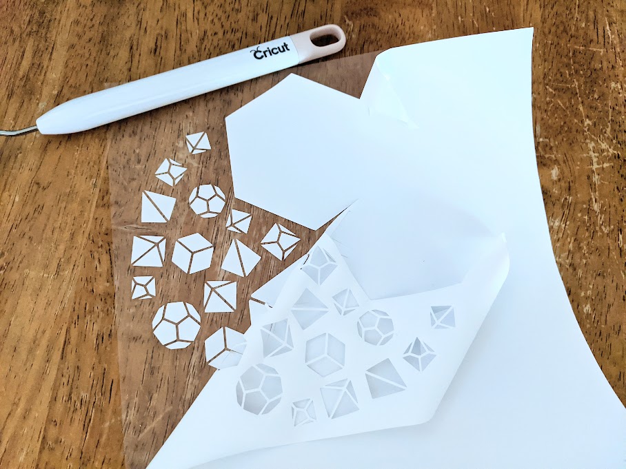 Overhead view of dice design cut on white iron on vinyl partially weeded. Weeding tool laying above design on table.