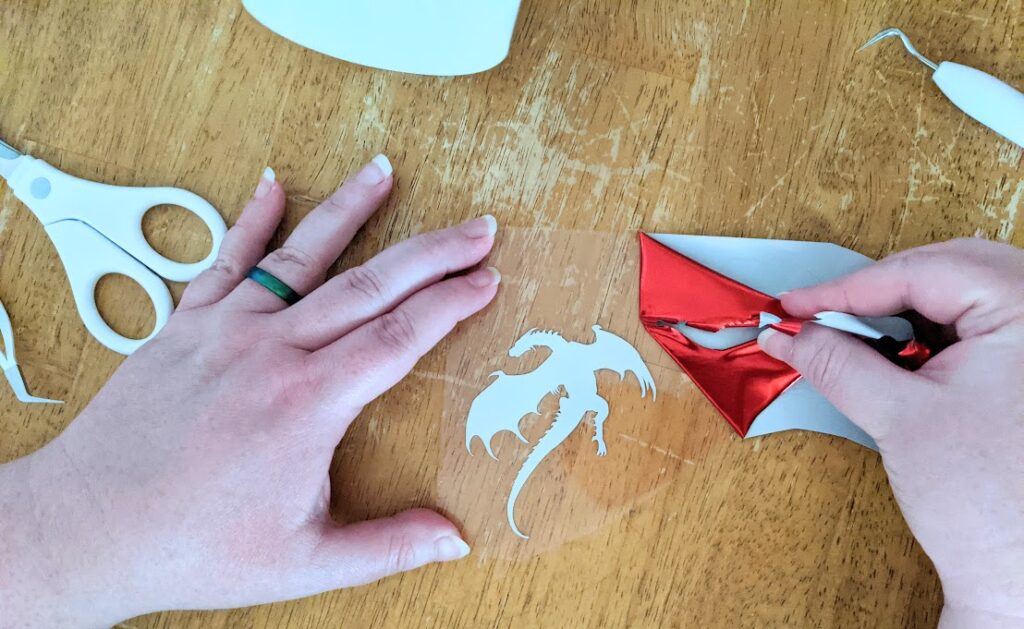 Overhead view of white woman's hands holding iron-on vinyl liner in place while pulling back excess red foil iron on revealing dragon silhouette design.