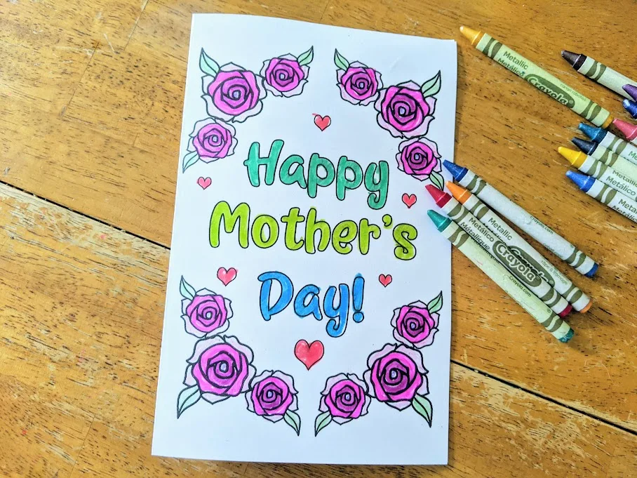 Colored in Mother's Day card laying on table with crayons laying next to it. Card says Happy Mother's Day! on front and has roses and hearts around it.