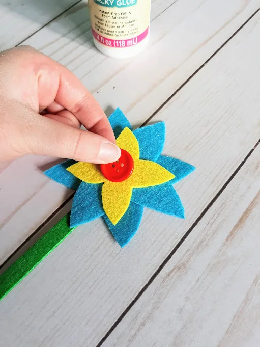 White woman's hand gluing red button to center of yellow and blue felt flower.