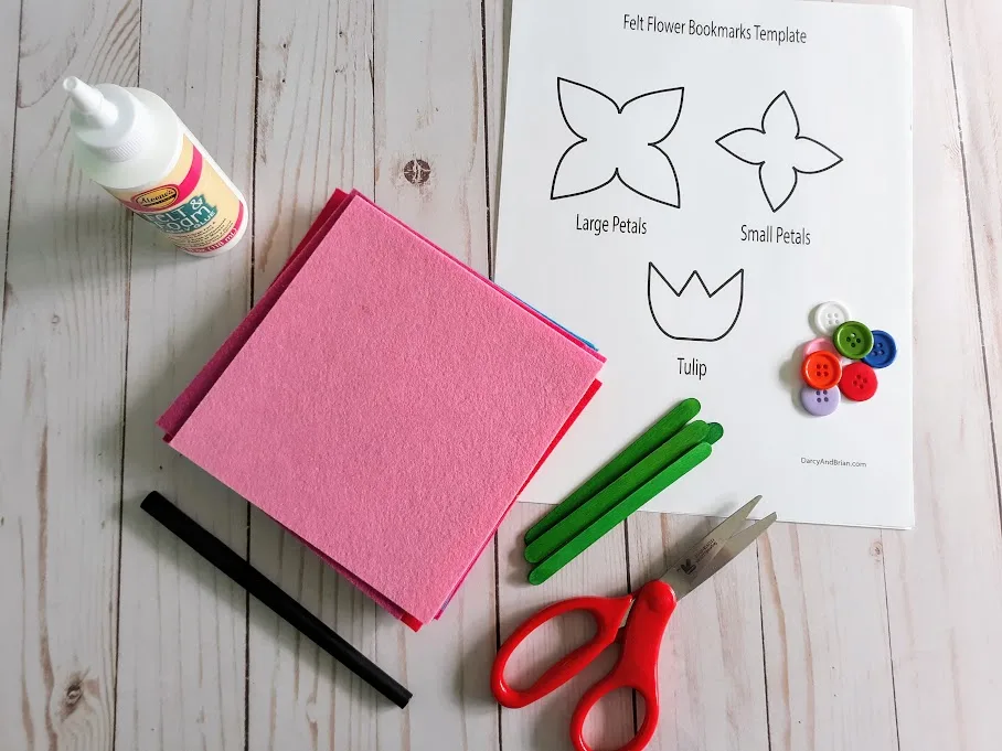 Overhead view of flower template printed out, pile of different colored buttons, green popsicle craft sticks, small red handled scissors, stack of felt squares, and felt tacky glue.