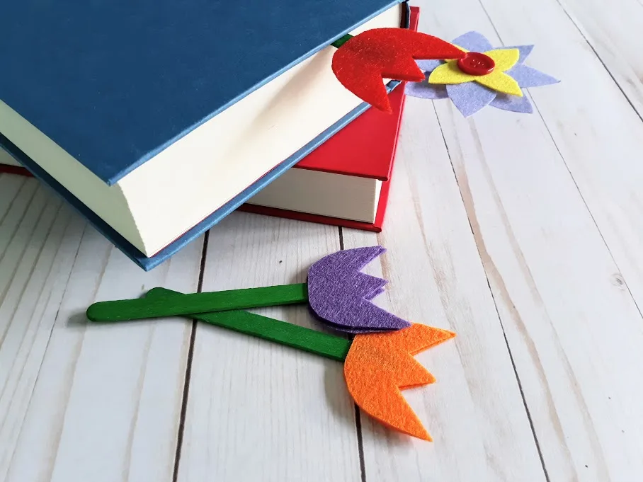 Blue and red hardcover books without dust jackets stacked on top of one another. Multi-petal felt flower bookmark sticking out of bottom book and red felt tulip bookmark sticking out of the top book. Purple and orange tulip bookmarks on green popsicle sticks lay next to the books.