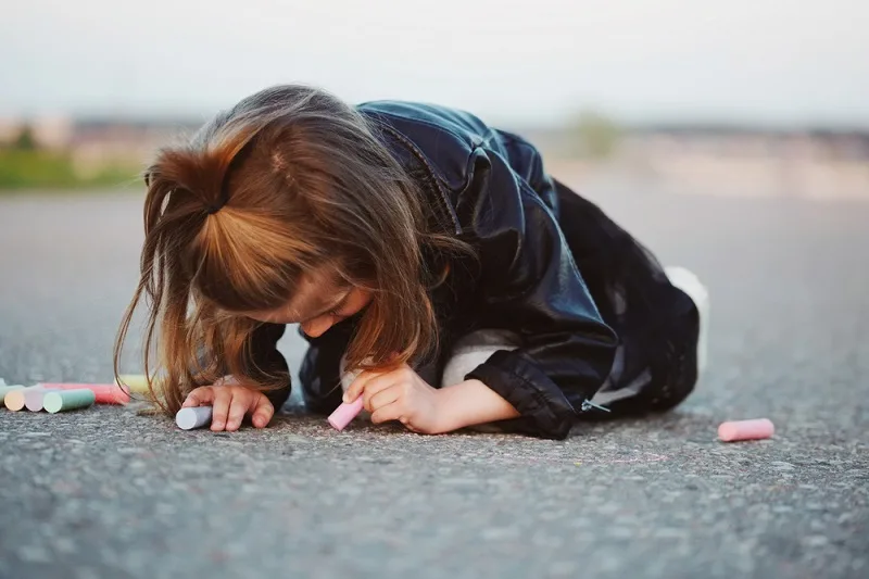 Young white girl with hair in partial ponytail drawing with sidewalk chalk outside.