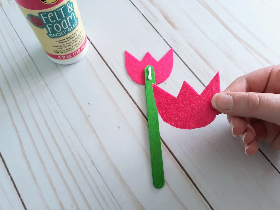 White woman's hand gluing green popsicle stick to pink felt tulip shape and gluing another felt piece to the other side.
