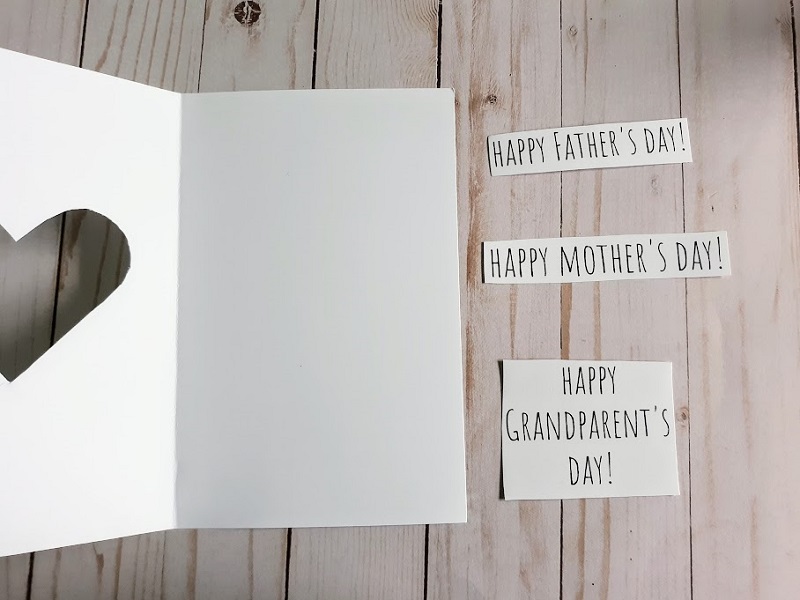 White cardstock paper folded in half with heart cut out laying open on table next to cut out phrases that read Happy Father's Day! Happy Mother's Day! Happy Grandparent's Day!