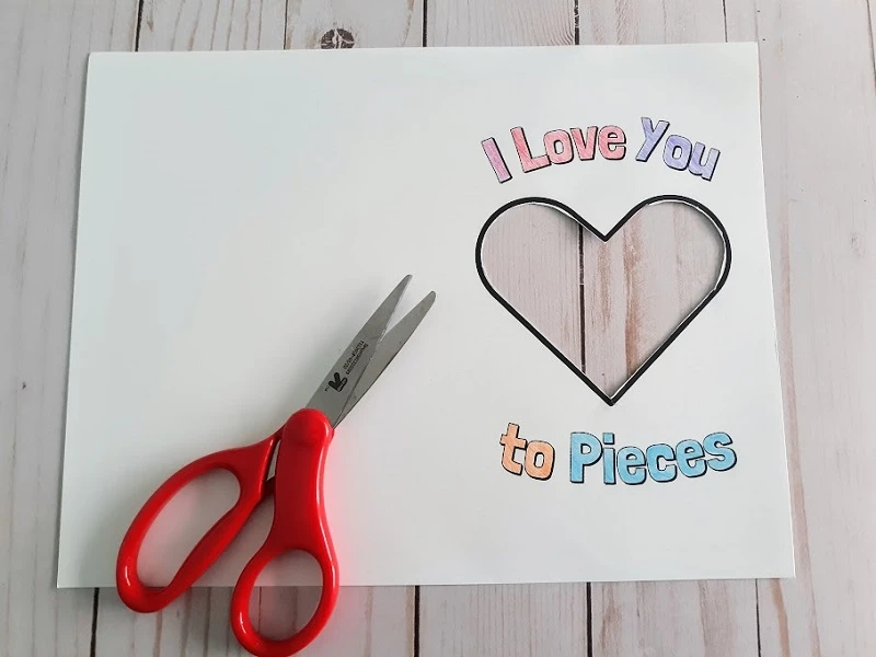 Overhead view of printed out card with center heart cut out. I Love You To Pieces words are colored in with colored pencil. Small pair of scissors laying over the paper.