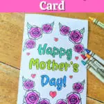 Top has white text outlined with black on a bright pink rectangle. Text reads Printable Mother's Day Card. Below text is a printed out card that says Happy Mother's Day colored in with blue and green marker. Words are surrounded by roses and small hearts colored in with crayons. Several crayons lay by card on table.