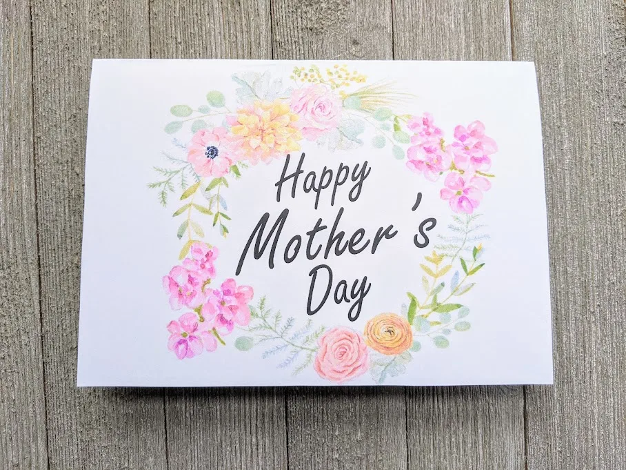 Overhead view of printable Mother's Day card printed out and folded. Front of card has a wreath of pink, yellow, and orange flowers with Happy Mother's Day in the center.