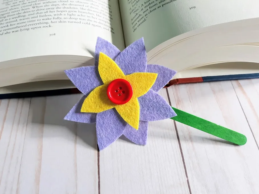 Purple and yellow felt flower with red button in the center attached to a green popsicle stick leaning against an open hardcover book.