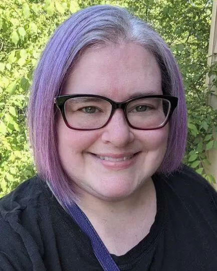 Photograph of blog author, Darcy. White woman with a light purple bob haircut and purple glasses standing in front of a green bush.