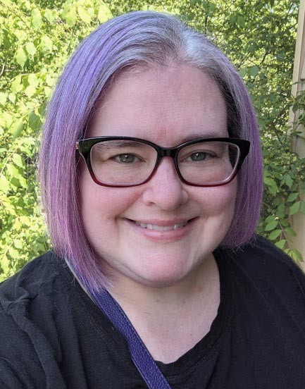 Headshot photo of site owner Darcy, a white woman with light purple and gray hair, smiling, wearing glasses, in a black shirt in front of a green bush.