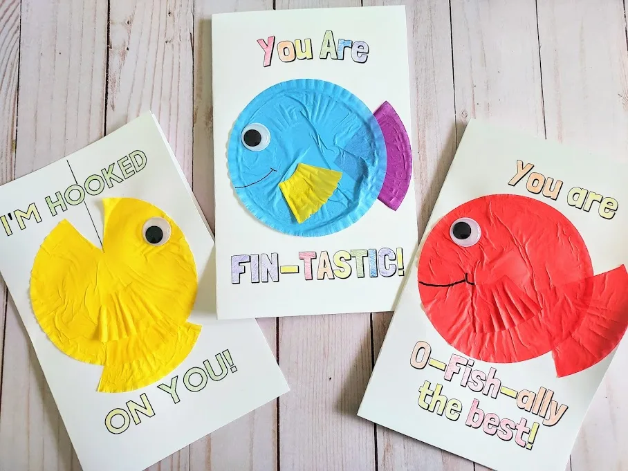 Overhead view of three completed fish pun card crafts. Three different printable cards with cupcake liner fish crafts on them. Left card says I'm Hooked On You with a yellow cupcake liner fish. Middle card says You Are Fin-Tastic with a blue, purple, and yellow cupcake liner fish. Right card says You Are O-fish-ally the best with a red cupcake liner fish.