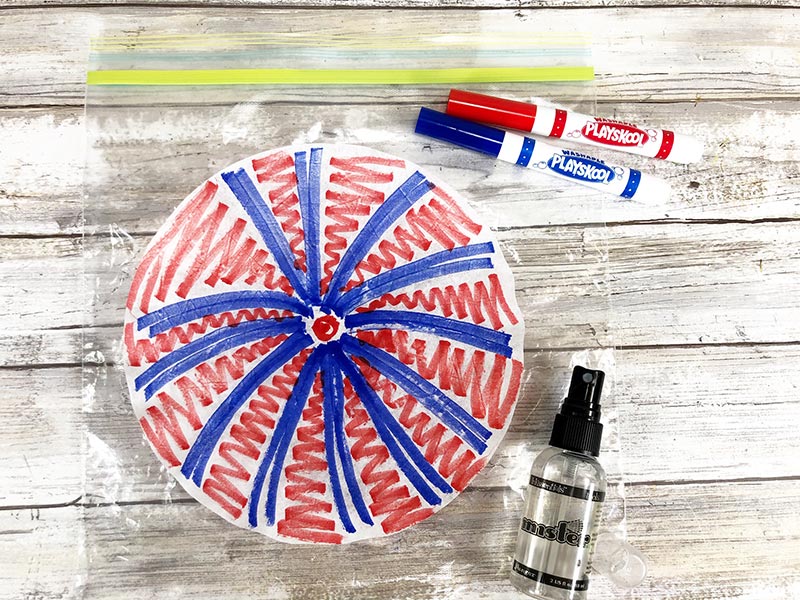 Coffee filter laying on a ziptop bag and colored with blue starburst lines and red marker between blue lines. Markers and small spray bottle next to it.