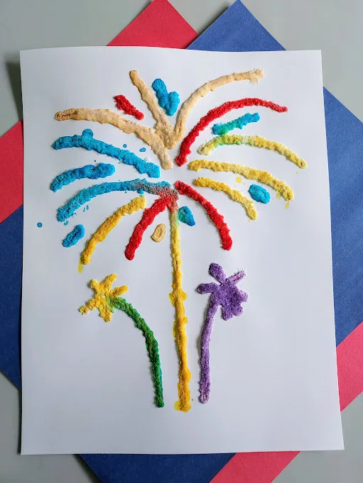 Overhead view of bright and colorful salt painting of fireworks using printable template. White paper with multicolored painted salt with red and blue construction paper underneath.