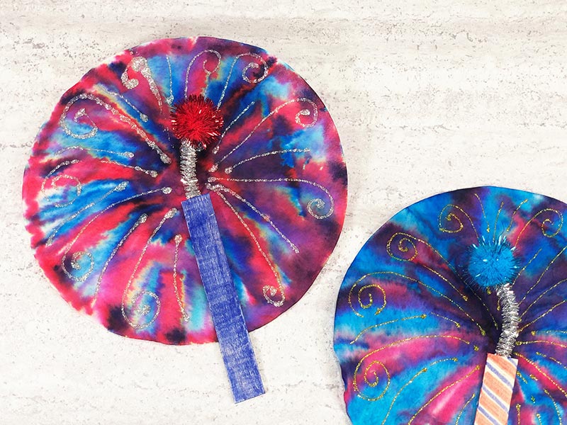 Overhead close up view of red and blue coffee filter fireworks with blue craft stick. To the right side is part of a mostly blue coffee filter firework too.