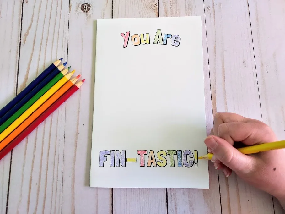 Overhead view of printable card that says You Are Fin-Tastic with colored pencils laying next to it. White girl's hand holding a color pencil and coloring in the words on the card.
